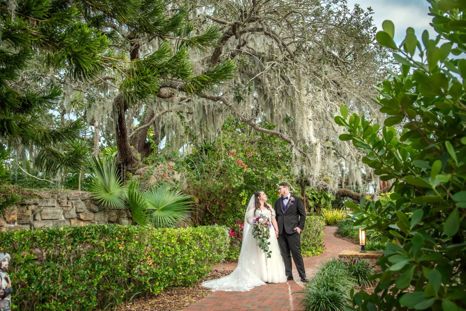 Newlyweds beneath sweeping oak branches on a brick garden path