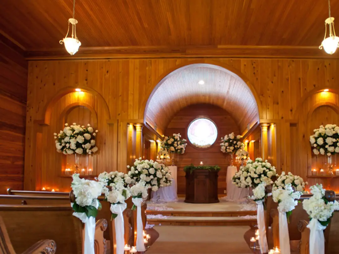 Wood-paneled chapel interior with white floral arrangements lining the aisle and altar