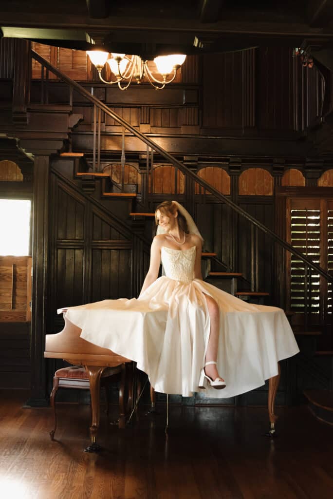 Bride seated by a piano and historic wooden staircase before her wedding
